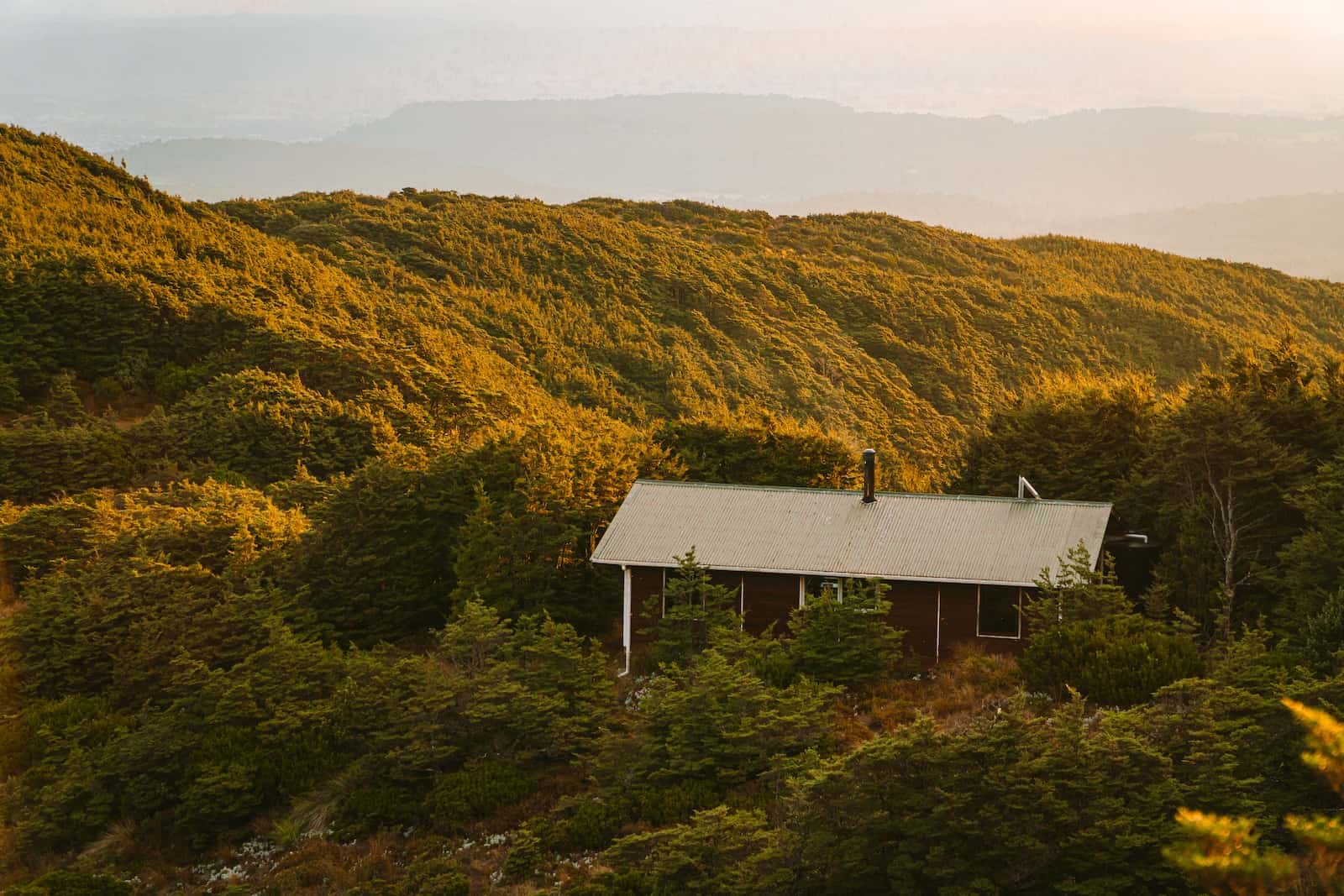 A backcountry hut on a bush-covered ridge at sunset, with ranges fading into the distance
